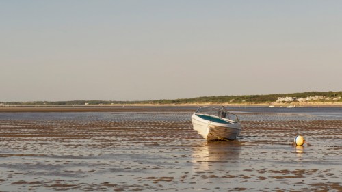 A small boat stranded on a sandy shore at low tide, with a buoy nearby and a distant shoreline in the background.