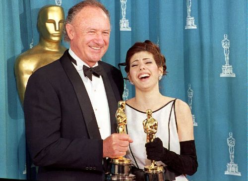 A man and a woman in formal attire smile and hold Oscars, standing in front of an Oscar backdrop.