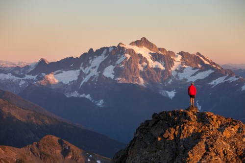 A person in a red jacket stands on a rocky outcrop, overlooking a majestic snow-capped mountain at sunset.