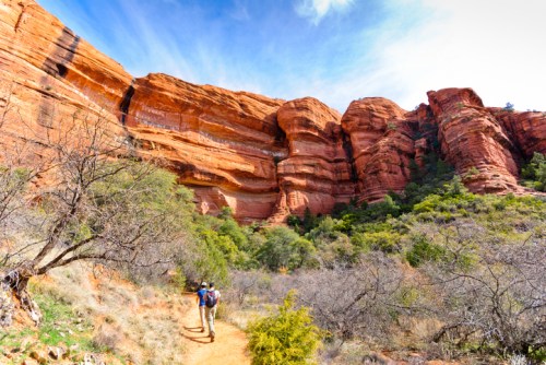 Two hikers walk along a trail surrounded by towering red rock formations and lush greenery under a blue sky.