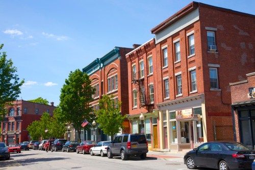 A sunny street lined with brick buildings and parked cars, featuring trees and an American flag in the background.
