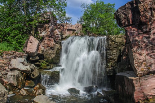 A serene waterfall cascades over rocky terrain, surrounded by lush greenery and a clear blue sky.