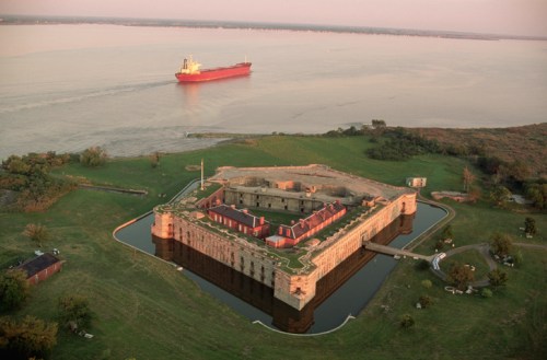 Aerial view of a historic fort surrounded by water, with a cargo ship passing in the background during sunset.