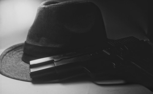A black and white image of a fedora hat resting beside a handgun on a flat surface.