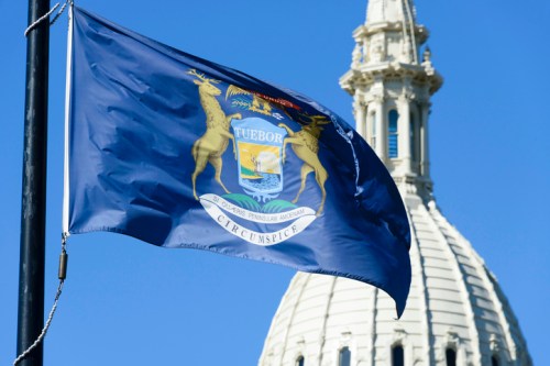 Michigan state flag waving in front of a dome structure against a clear blue sky.
