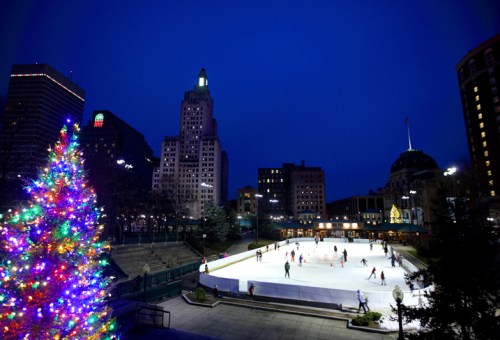 A vibrant ice skating rink surrounded by city buildings, with a decorated Christmas tree in the foreground at dusk.