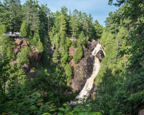 A scenic waterfall cascading down rocky cliffs, surrounded by lush green trees and a viewing platform above.