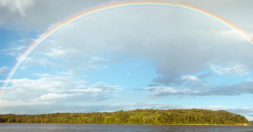A vibrant rainbow arcs over a lush green island, with a blue sky and scattered clouds in the background.