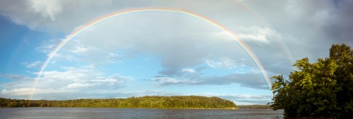 A vibrant rainbow arcs over a serene lake, surrounded by lush greenery and a partly cloudy sky.
