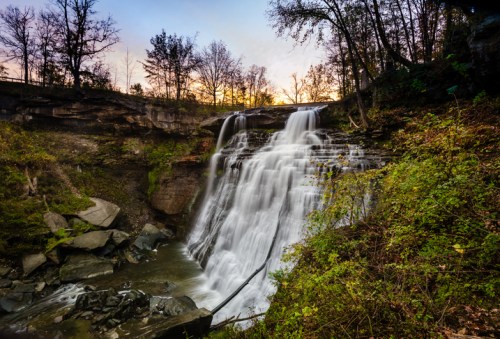 A serene waterfall cascades over rocky ledges, surrounded by trees and bathed in warm sunset light.