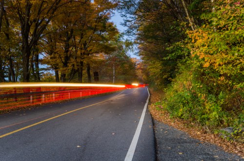 A winding road lined with colorful autumn trees, featuring light trails from passing cars at dusk.