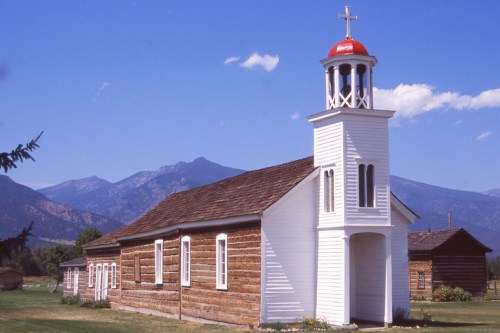 A white church with a red roof and cross, set against a backdrop of mountains and blue sky.