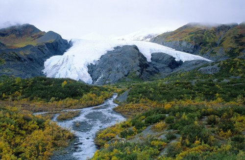 A glacier flows down rocky terrain, surrounded by autumn foliage and a winding stream under a cloudy sky.