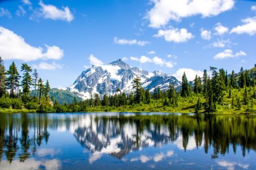 A serene lake reflects a snow-capped mountain and lush green trees under a bright blue sky with fluffy clouds.