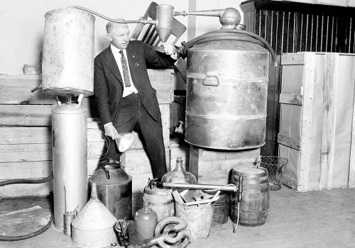 A man in a suit stands beside various metal distilling equipment and containers, showcasing a vintage setup.
