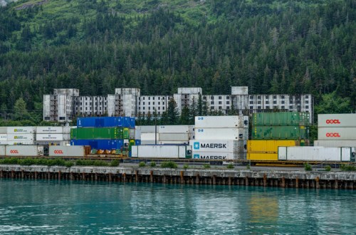 A stack of colorful shipping containers by a calm waterway, with a dilapidated building and forested hills in the background.