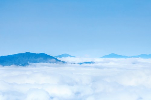 A serene landscape of mountains partially obscured by a blanket of fluffy white clouds under a clear blue sky.