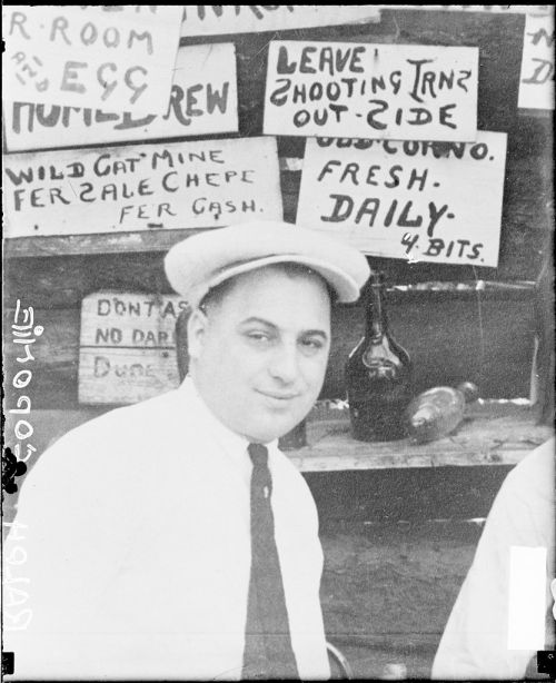 A man in a hat sits in front of various signs, including "Leave Shooting Trans Outside" and "Wild Cat Mine for Sale."