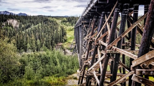 A wooden trestle bridge stands over lush green trees and hills under a cloudy sky.