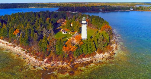 Aerial view of a lighthouse on a lush, green island surrounded by clear blue water and autumn-colored trees.