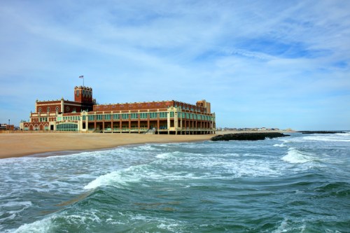 Historic building by the beach with waves crashing in the foreground and a cloudy sky above.
