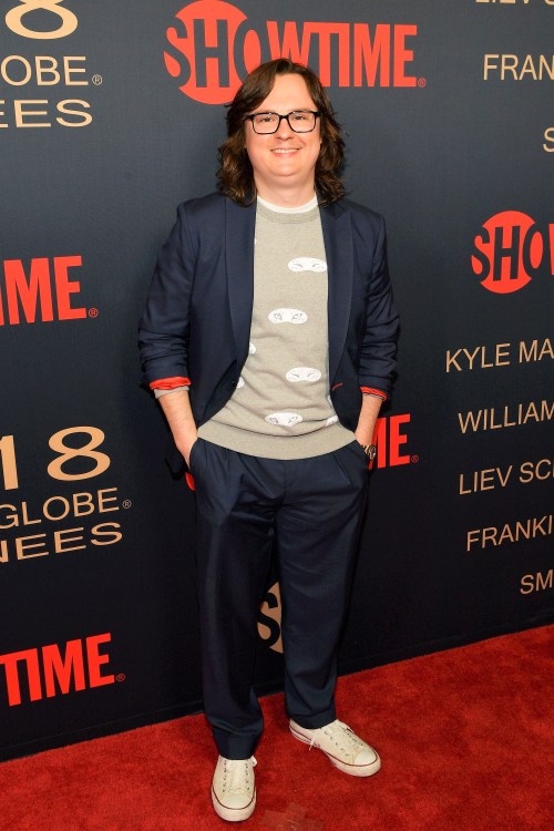 A person in a gray sweater and navy suit stands on a red carpet with a Showtime backdrop, smiling confidently.