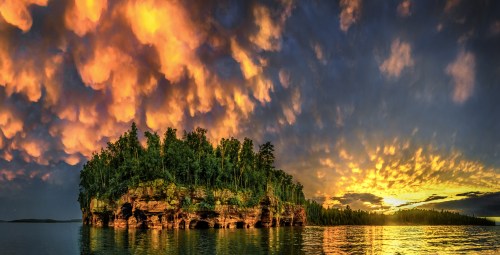 A vibrant sunset over a lush island, surrounded by calm waters and dramatic, colorful clouds in the sky.