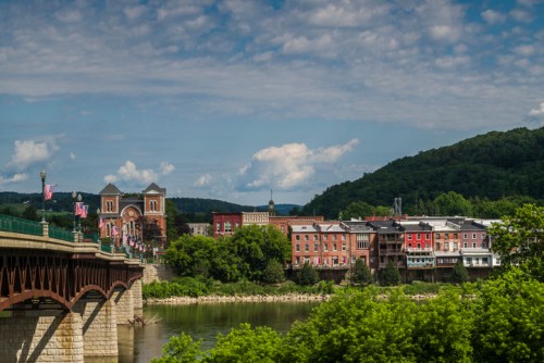 A scenic view of a riverside town with colorful buildings, a bridge, and lush green hills under a partly cloudy sky.