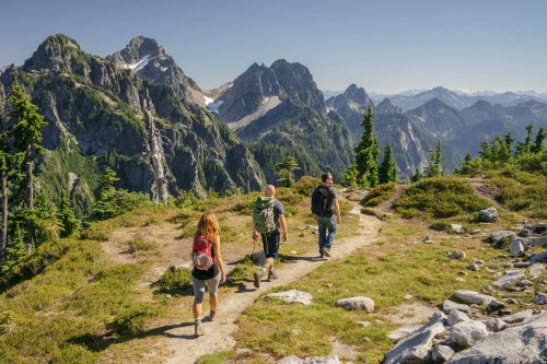 Three hikers walking along a trail in a mountainous landscape with lush greenery and distant peaks under a clear blue sky.
