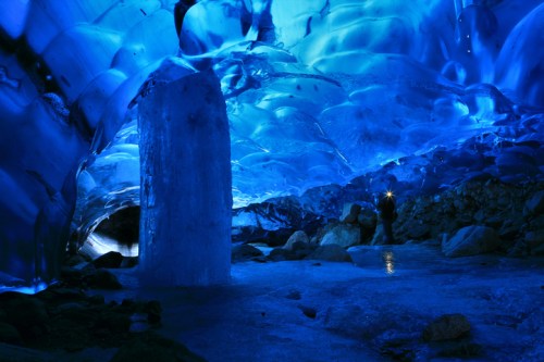 A stunning ice cave illuminated in blue, featuring icy formations and a person with a flashlight exploring the scene.