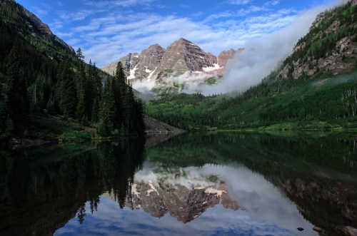 A serene lake reflects towering mountains and lush green trees under a blue sky with scattered clouds.