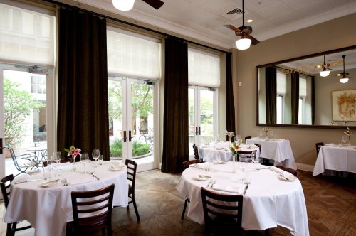 Elegant restaurant interior with white tablecloths, wooden chairs, and large windows overlooking a garden.