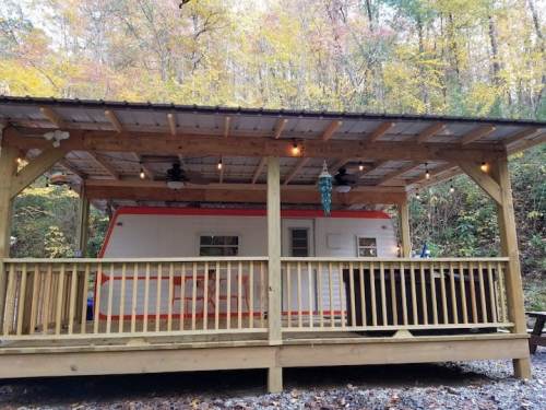 A vintage camper on a wooden deck surrounded by autumn trees, with lights and fans under a covered roof.