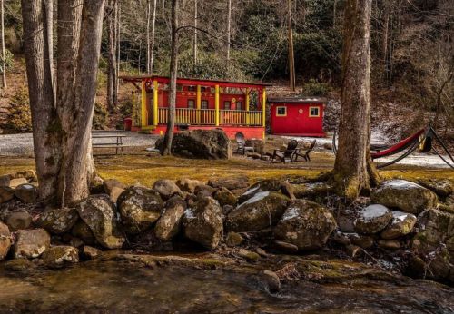 A colorful cabin by a stream, surrounded by trees and rocks, with a hammock and seating area nearby.