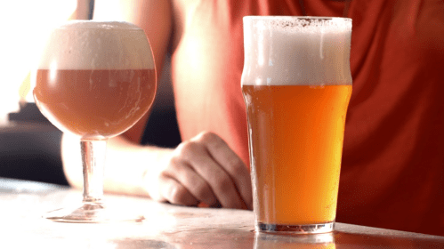 Two glasses of beer on a bar: one in a stemmed glass with a foamy top, the other in a pint glass, both golden in color.
