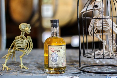 A bottle of Hamilton Smoke whiskey beside a skeleton figurine and a caged skeleton bird on a rustic surface.