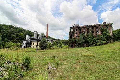 Abandoned industrial buildings overgrown with vegetation, set against a cloudy sky and green landscape.