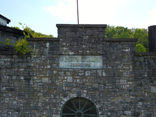 Stone wall of a distillery with a sign reading "L. E. B. R. Y. Distillery, Founded 1884," surrounded by greenery.