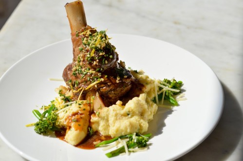 A plated dish featuring a braised lamb shank on creamy mashed potatoes, garnished with herbs and vegetables.