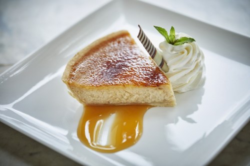 A slice of creamy dessert with a caramelized top, drizzled with sauce, and accompanied by whipped cream and mint.
