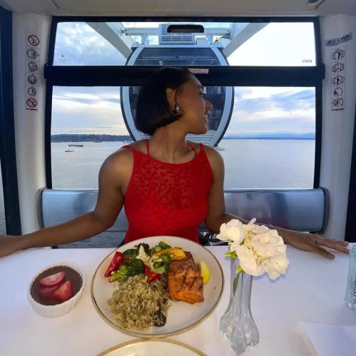 A woman in a red dress sits at a table with a meal, enjoying a scenic view from a ferris wheel cabin.