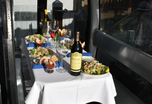 A table set with gourmet salads, shrimp, wine, and cocktails, inside a cozy gondola with rain on the windows.