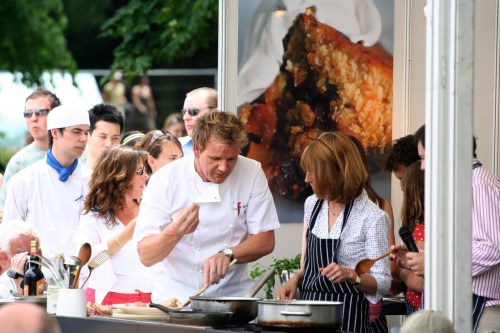 A chef demonstrates cooking at an outdoor event, surrounded by an audience and fellow cooks.