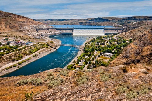 A scenic view of a river valley with a dam, bridge, and a small town surrounded by hills and greenery.
