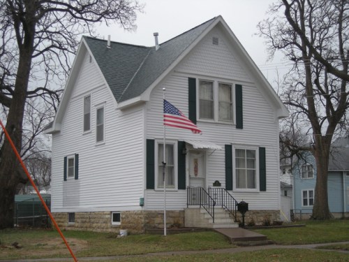 A white two-story house with green shutters and an American flag in front, surrounded by bare trees.
