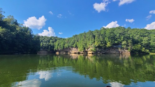 A serene lake surrounded by lush green trees and rocky cliffs under a bright blue sky with fluffy white clouds.