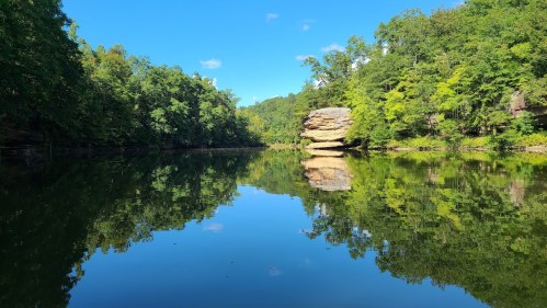 A serene river scene with lush green trees and a large rock formation reflected in the calm water under a clear blue sky.
