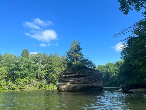 A large rock formation with a tree on top, surrounded by calm water and lush green trees under a clear blue sky.
