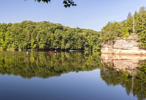 A serene lake surrounded by lush green trees and a rocky cliff, with a small boat gliding across the calm water.