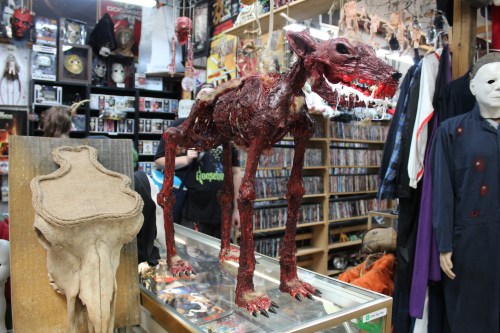 A grotesque, skeletal dog sculpture stands on display in a horror-themed shop, surrounded by various horror memorabilia.
